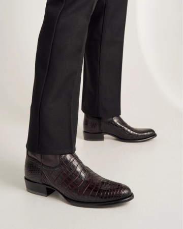 Person wearing black dress pants and dark brown, crocodile-patterned leather ankle boots standing on a white surface.