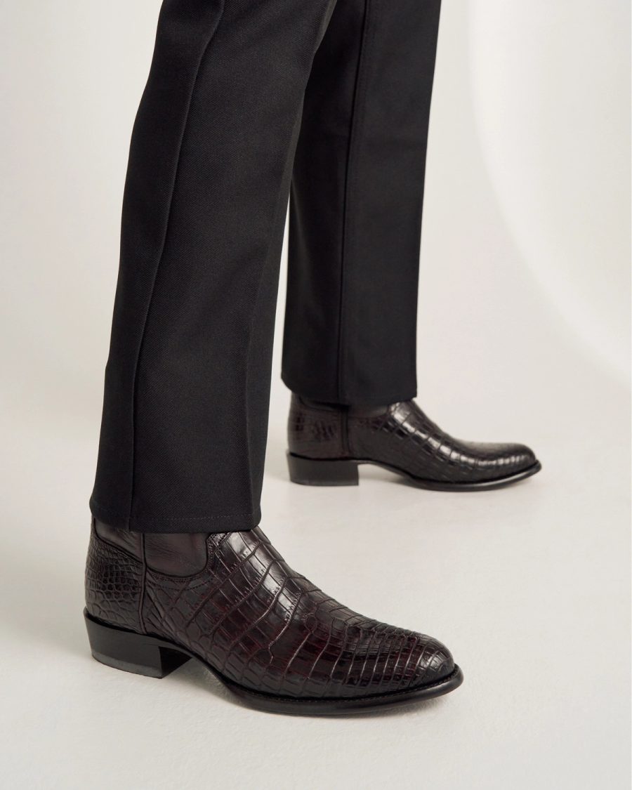 Person wearing black dress pants and dark brown, crocodile-patterned leather ankle boots standing on a white surface.