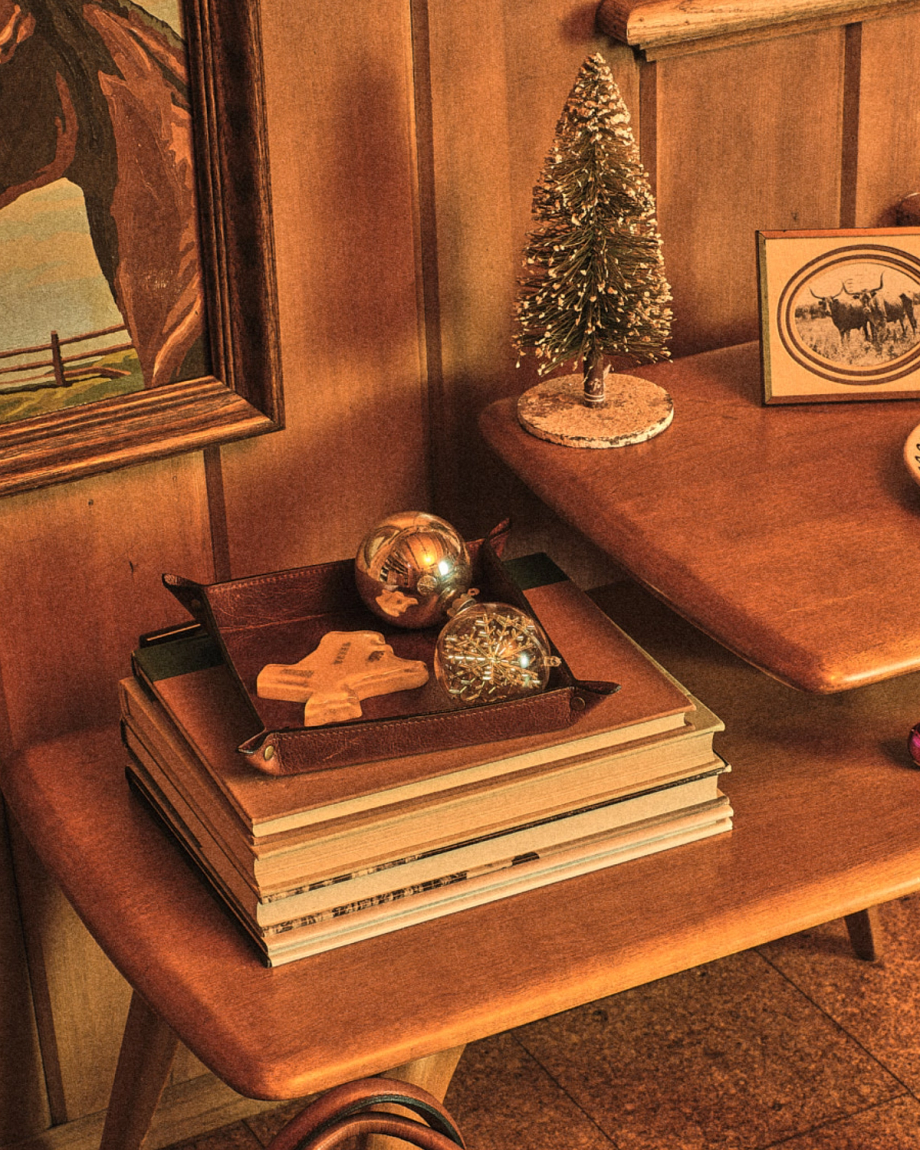A wooden table with a stack of books, a leather tray holding two ornaments and a cookie, and a small decorative Christmas tree in a vintage-styled room.