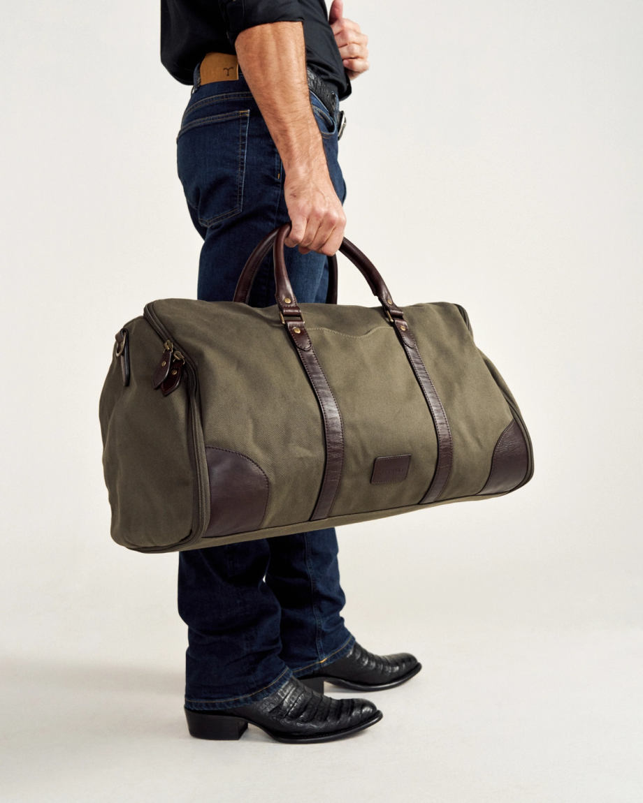 Person holding a large olive green and brown duffel bag, wearing blue jeans and black shoes.