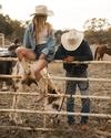 A person sits on a fence near a horse, wearing a cowboy hat, denim jacket, and boots. Another person leans on the fence, also in a cowboy hat, beside a grazing horse.