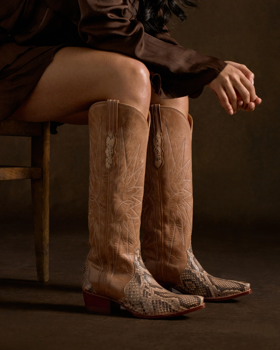 The Loretta in Mangrove Invasive Python. A person sitting on a wooden chair, wearing a brown dress and tall tan western cowboy boots with a snake-skin pattern inspired by the Loretta Invasive Python.