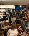 People shop and socialize inside a well-lit clothing and accessories store, with some seated and others browsing or standing in groups.