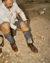 A person in work clothes sits on a rough, gritty surface, holding knee pads, with some tools visible nearby.