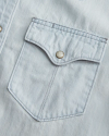 Close-up of a light blue denim shirt pocket with a buttoned flap, showing stitching details.