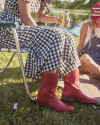 Two people are sitting outdoors. One wears a checkered dress with red boots, holding a drink can. The other wears a hat and overalls, sitting on the ground. Green grass and trees are in the background.