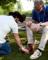 A man on one knee shines the brown leather shoes of a seated man wearing beige pants and a blue shirt, outdoors on grass.