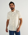 A man with short dark hair and a beard wears a light-colored, short-sleeve button-up shirt with two front pockets and blue jeans, standing against a plain white background.