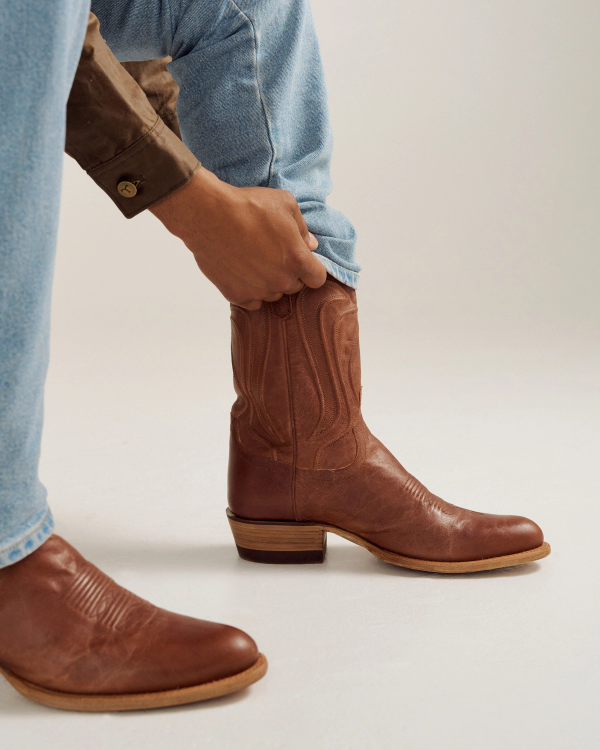 A person wearing light blue jeans and a brown shirt adjusts a brown leather cowboy boot on their left foot.