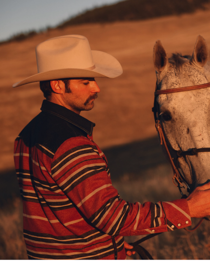 Cowboy in a striped jacket and hat stands beside a white horse in a sunlit open field.