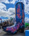 A large, colorful painted cowboy boot sculpture under a partly cloudy sky, with a person sitting nearby.