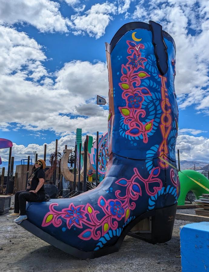 A large, colorful painted cowboy boot sculpture under a partly cloudy sky, with a person sitting nearby.