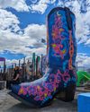 A large, colorful painted cowboy boot sculpture under a partly cloudy sky, with a person sitting nearby.