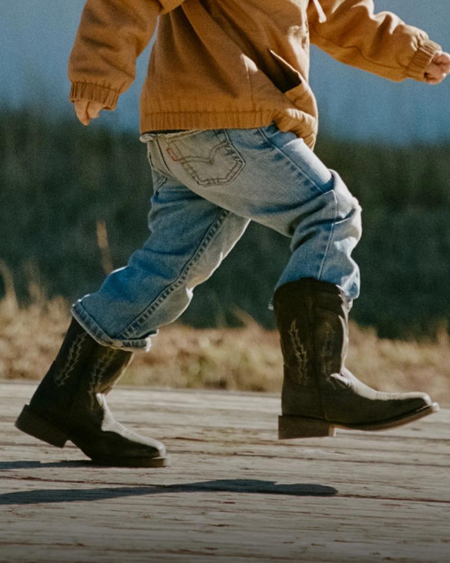 A child wearing a brown sweater, blue jeans, and black cowboy boots walks outdoors on a wooden surface.