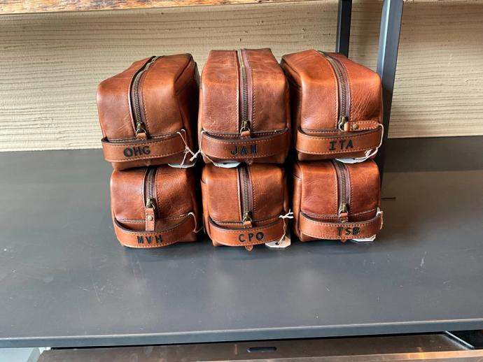 A stack of six brown leather dopp kits with zippers, each embroidered with different three-letter initials: OHG, JAM, ITA, MVH, CPO, and TSB.