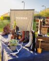 A display table with various cowboy boots, a succulent, and antlers in front of a Tecovas banner at an outdoor event.