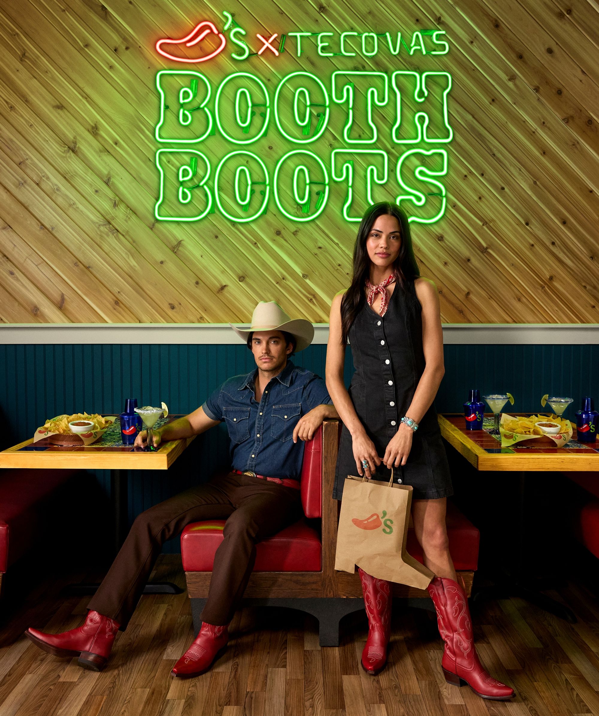 A man and woman in red cowboy boots pose in a restaurant booth beneath a neon sign that reads "SXT TECOVAS BOOTH BOOTS"; the woman holds a Chili’s takeout bag.