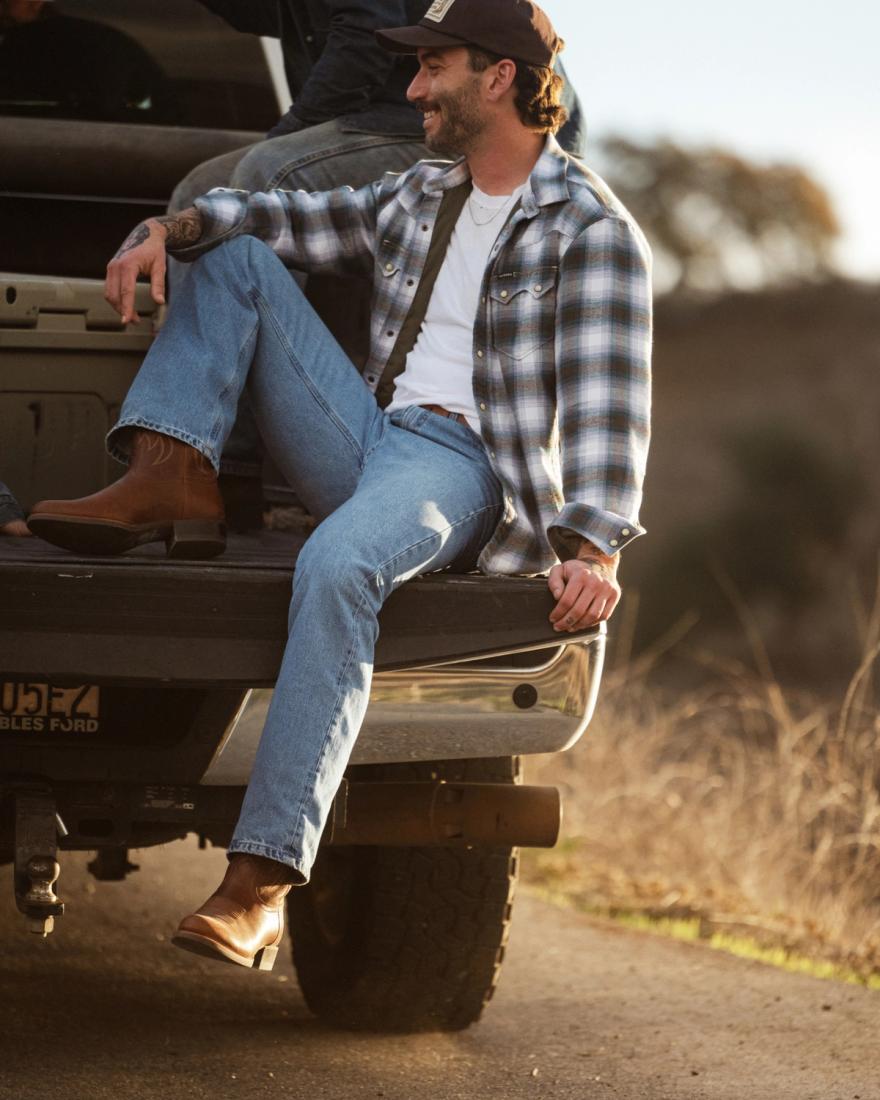A person in a plaid shirt and jeans sits on a truck tailgate outdoors, with another person partially visible. Trees and grass are in the background.