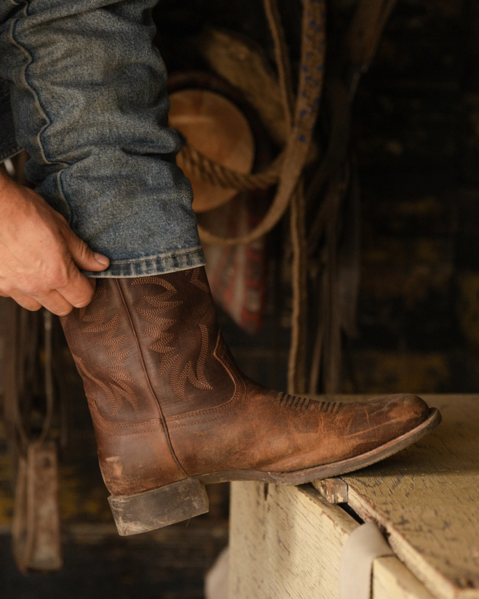 A person wearing blue jeans is pulling on a brown cowboy boot. A hat and ropes hang blurred in the background.