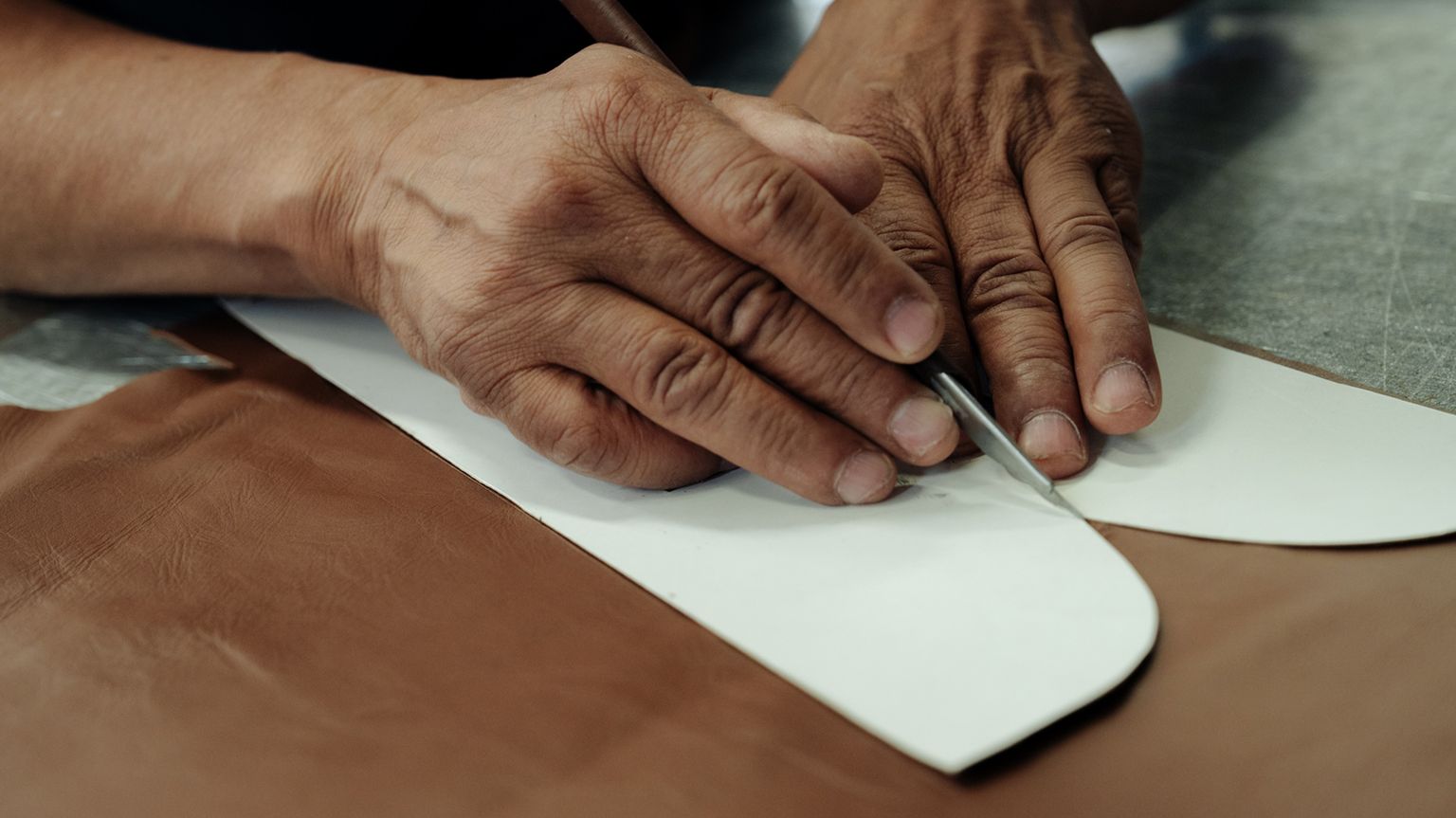 A person uses a cutting tool to trace around a paper pattern on a piece of brown leather on a work surface.
