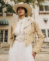 A person wearing a white dress, a beige fringed jacket, and a white cowboy hat stands outdoors in front of a building with striped awnings.