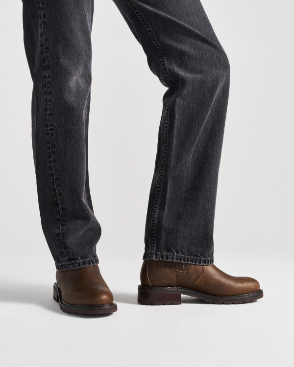 The Chelsea in Cafe Cowhide. Person wearing dark jeans and brown leather Cafe Cowhide shoes, standing on a white background. Only legs and feet are visible.