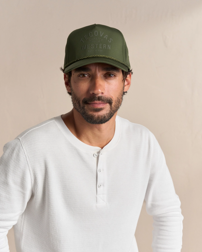 Man in a green baseball cap and white long-sleeve henley shirt stands against a neutral background.