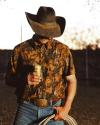 Man in a cowboy hat and western shirt holds a canned drink in one hand and a rope in the other, standing outdoors at sunset.