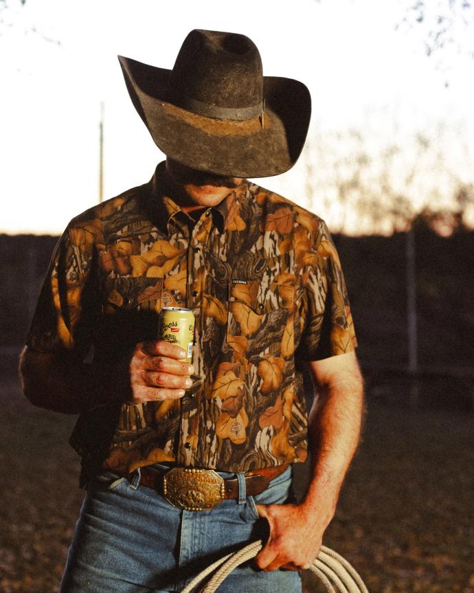 Man in a cowboy hat and western shirt holds a canned drink in one hand and a rope in the other, standing outdoors at sunset.