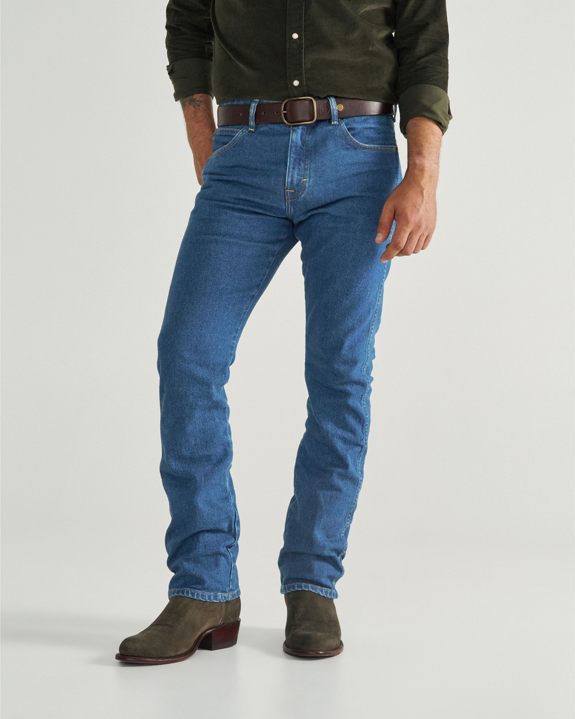 A person wearing a dark green shirt, blue jeans, a brown belt, and brown boots stands against a plain light background.