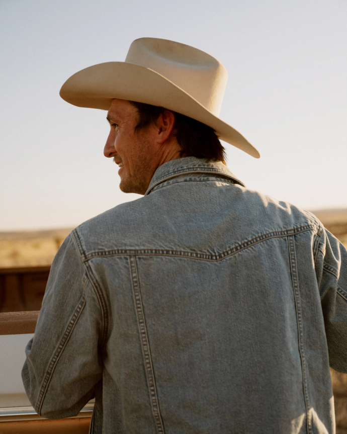 A man wearing a cowboy hat and denim jacket stands outdoors, facing away and looking toward an open, sunlit landscape.
