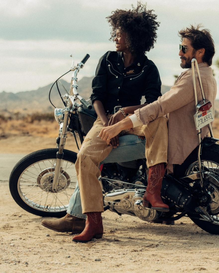 Two people sit on a motorcycle in a desert setting; the woman sits on the seat, while the man sits on the ground leaning against the bike.