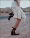 Person in a white dress lifts hem while wearing black western boots with a yellow floral design, standing on a dirt road in a desert landscape.