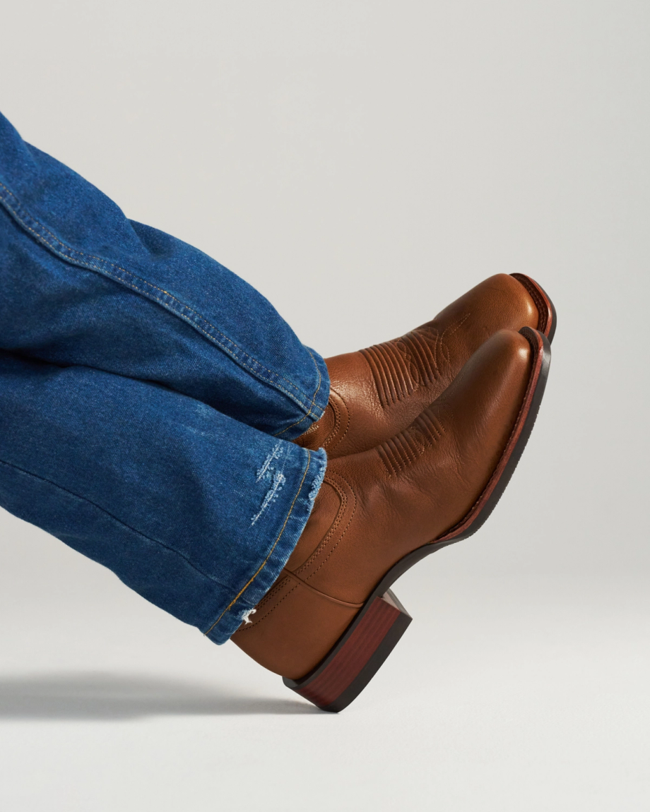 The Dusty in Mesa Goat. Person wearing blue jeans and brown leather boots with a low heel, sitting or reclining with legs crossed against a plain background, evokes the relaxed spirit of the Mesa Goat—rugged yet refined.