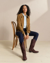 A person with long hair sits on a wooden chair, wearing a brown fringed jacket, a light blue shirt, dark jeans, and brown cowboy boots.