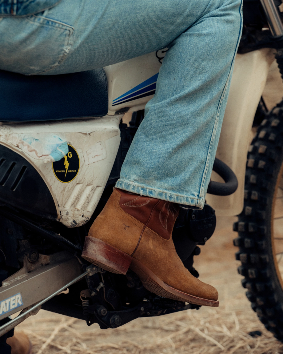 The Buck in Copper Cowhide. Person wearing jeans and brown cowhide boots sits on a white dirt bike, one foot resting on the footpeg, with dry grass scattered across the ground.