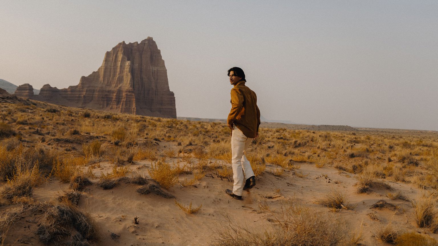 A man in a tan shirt and light-colored trousers walks through a desert landscape toward a large rock formation.