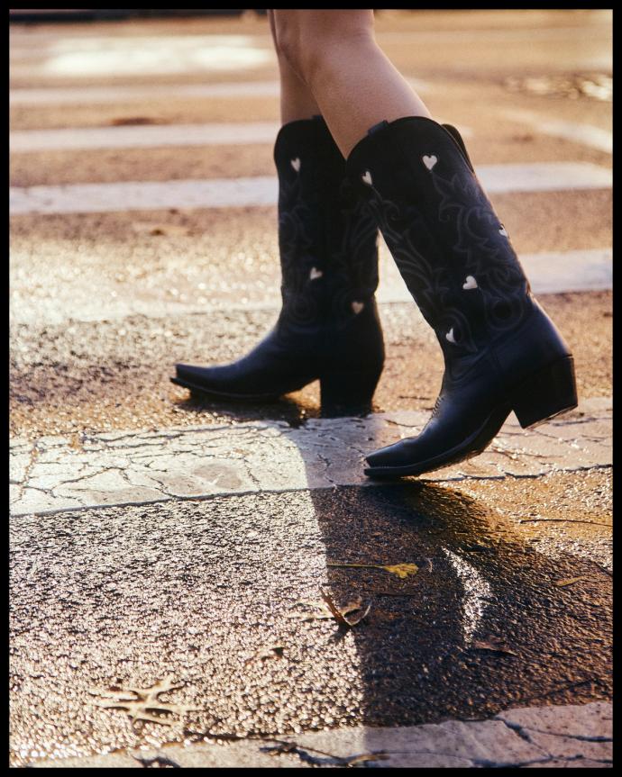 A person wearing black cowboy boots with white heart details crosses a street with a textured, wet surface.
