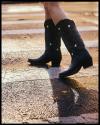 A person wearing black cowboy boots with white heart details crosses a street with a textured, wet surface.