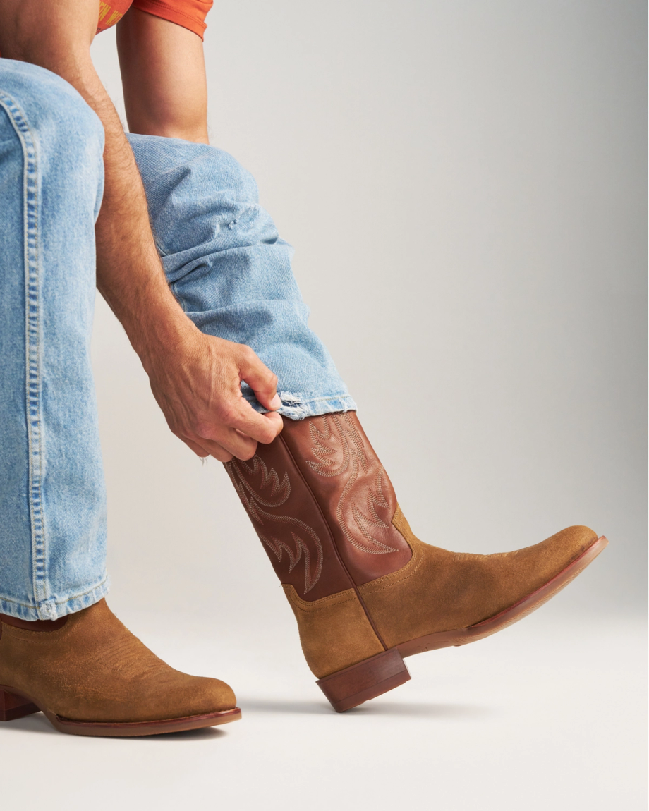 The Buck in Copper Cowhide. A person in blue jeans and an orange shirt tucks their pant leg into a sturdy Copper Cowhide cowboy boot.