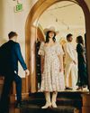A woman in a floral dress and cowboy hat stands indoors near an archway while three other people walk nearby.