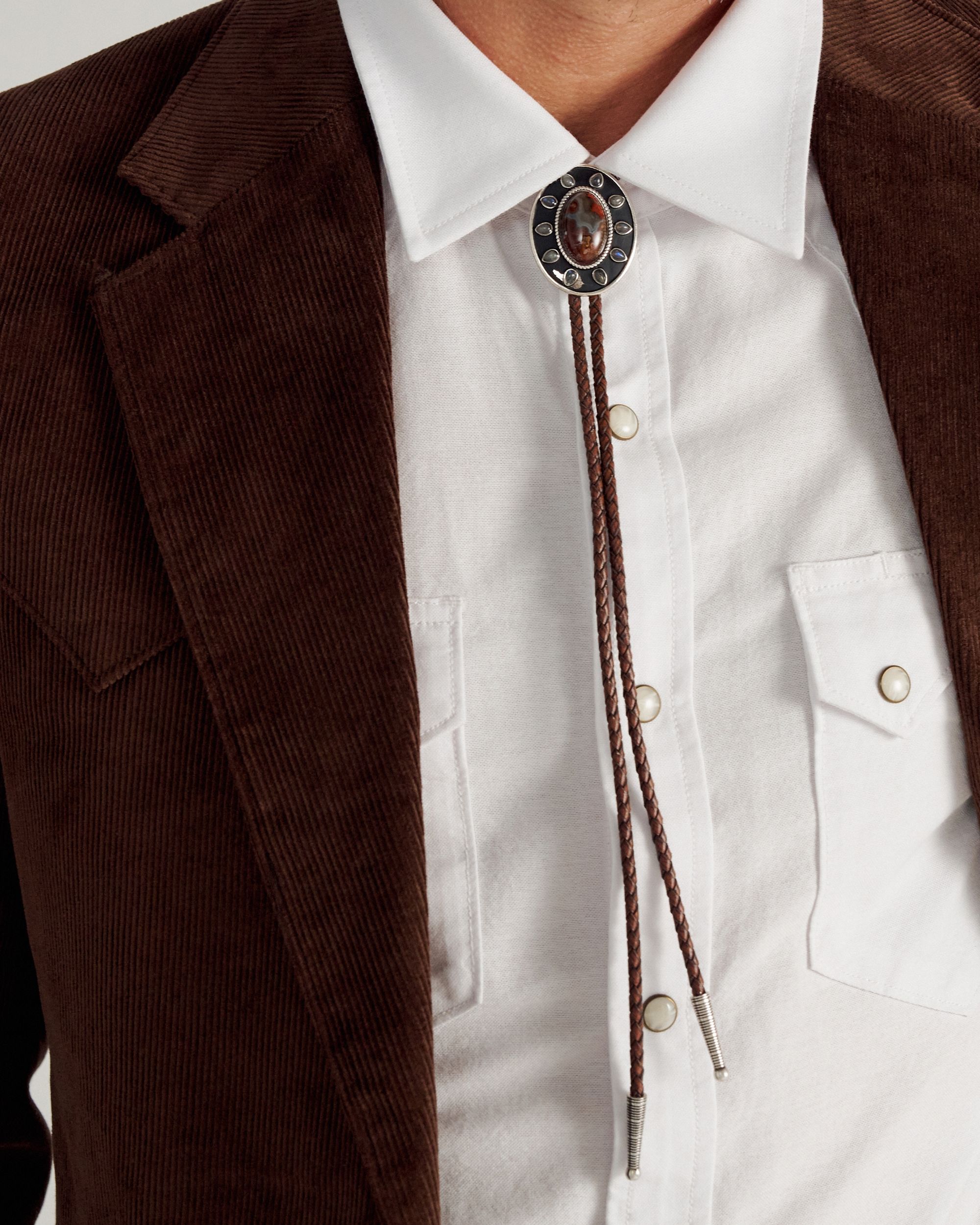 Close-up of a person wearing a white collared shirt, brown jacket, and a decorative bolo tie with a dark oval stone and metal accents.