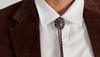 Close-up of a person wearing a white collared shirt, brown jacket, and a decorative bolo tie with a dark oval stone and metal accents.