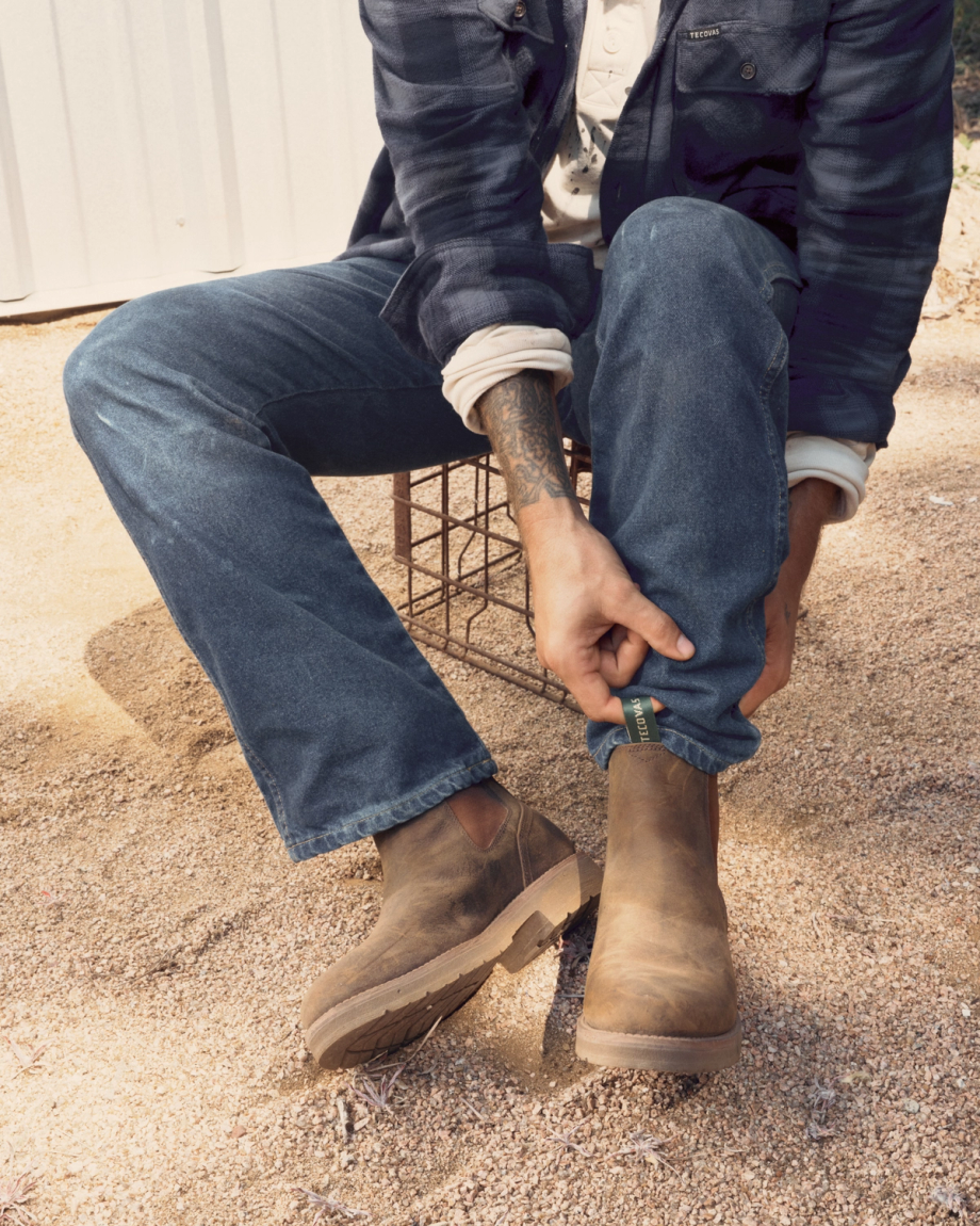 The Rugged Chelsea in Canteen Cowhide. A person sitting outdoors tucks the hem of their jeans into brown leather boots—The Rugged Chelsea Cowhide—with visible sand and a wire crate nearby.