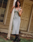 A woman in a light blue dress and The Sadie Hair on Hide / Cowhide boots leans against a white post in front of a beige wooden house.