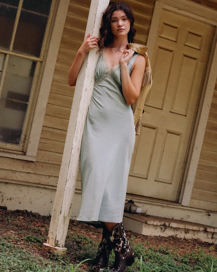A woman in a light blue dress and The Sadie Hair on Hide / Cowhide boots leans against a white post in front of a beige wooden house.