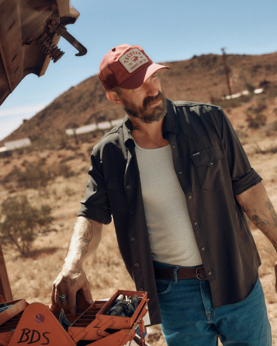 A bearded man in a red cap and unbuttoned shirt stands outdoors by an open car hood, with desert hills in the background.