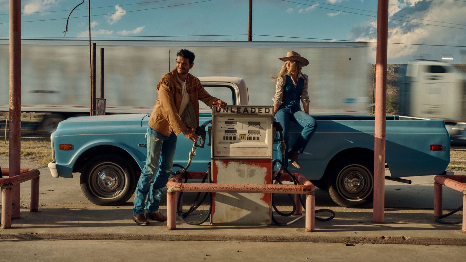A man and woman in Western attire stand by an old gas pump with a blue vintage pickup truck; a blurred semi-truck passes by in the background.