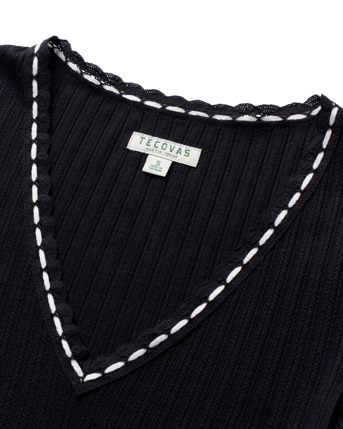 Close-up of a black V-neck ribbed fabric with white crochet trim, featuring a Tecovas label indicating size small.