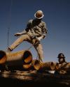 Two construction workers in hard hats handle large, rusty metal pipes with chains under a clear blue sky.